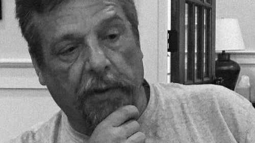 Black and white image of John Barnett with a beard, thoughtfully touching his chin, sitting in a room with shelves in the background