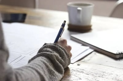 The girl writes at the table with a blue pen, and there is a cup next to it