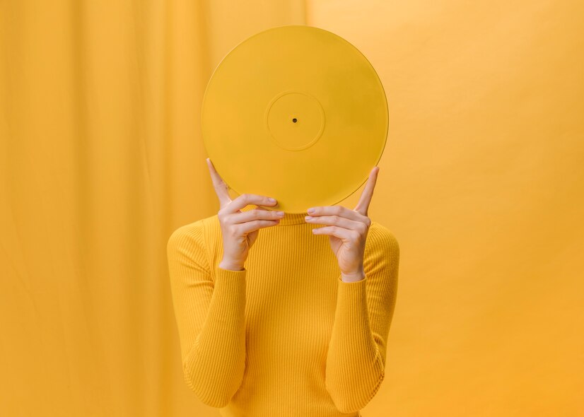 Жінка тримає в руках вінілову жовту платівку та закриває нею своє обличчя на жовтому фоні